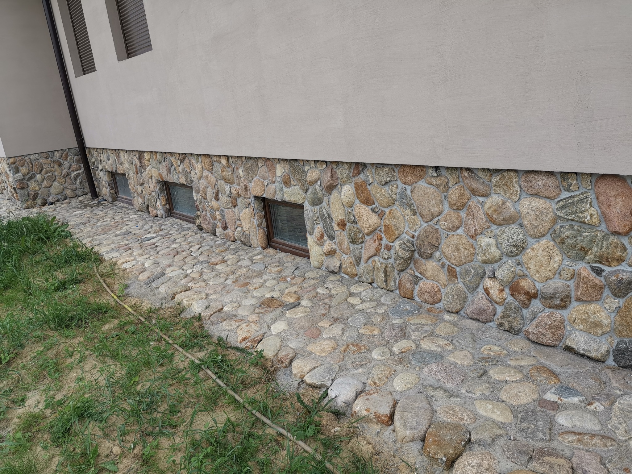 Wykończenie elewacji kamieniem rzecznym i ułożona z niego ścieżka wokół domu. Widoczne okna piwniczne i fragment trawnika. Kamień w różnych odcieniach szarości i beżu.