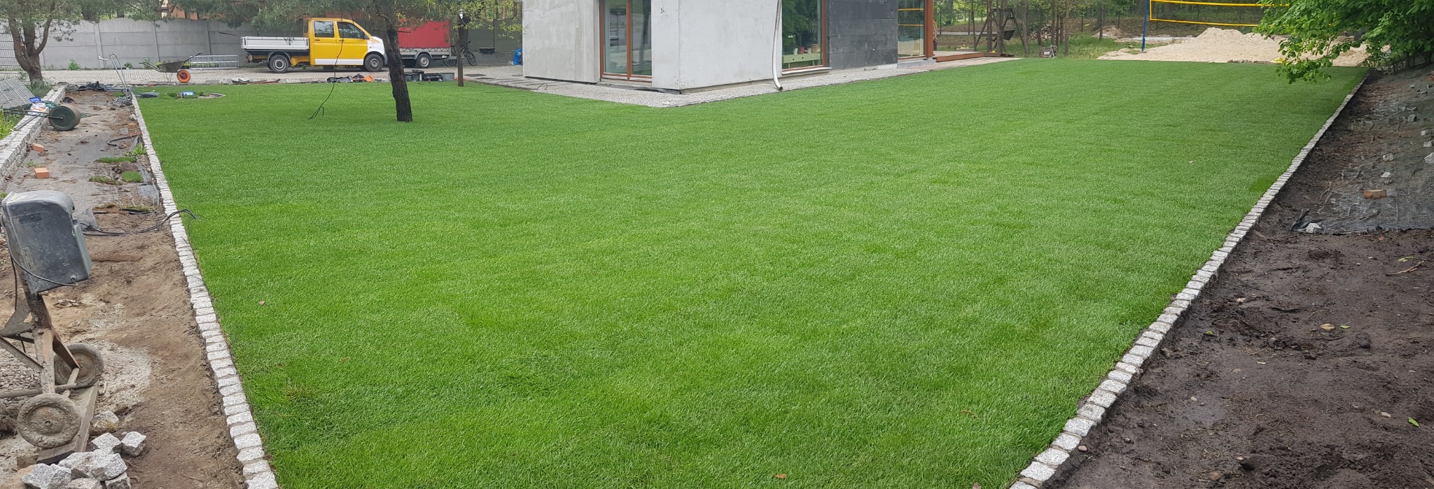 Rozległy trawnik z obrzeżami z kostki granitowej, fragment terenu w trakcie prac wykończeniowych. W tle budynek i żółty samochód dostawczy.