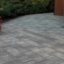 Ułożona kostka brukowa w odcieniach szarości na podjeździe, z zielenią w tle i fragmentem ceglanego muru, tworzy estetyczną przestrzeń przed domem.