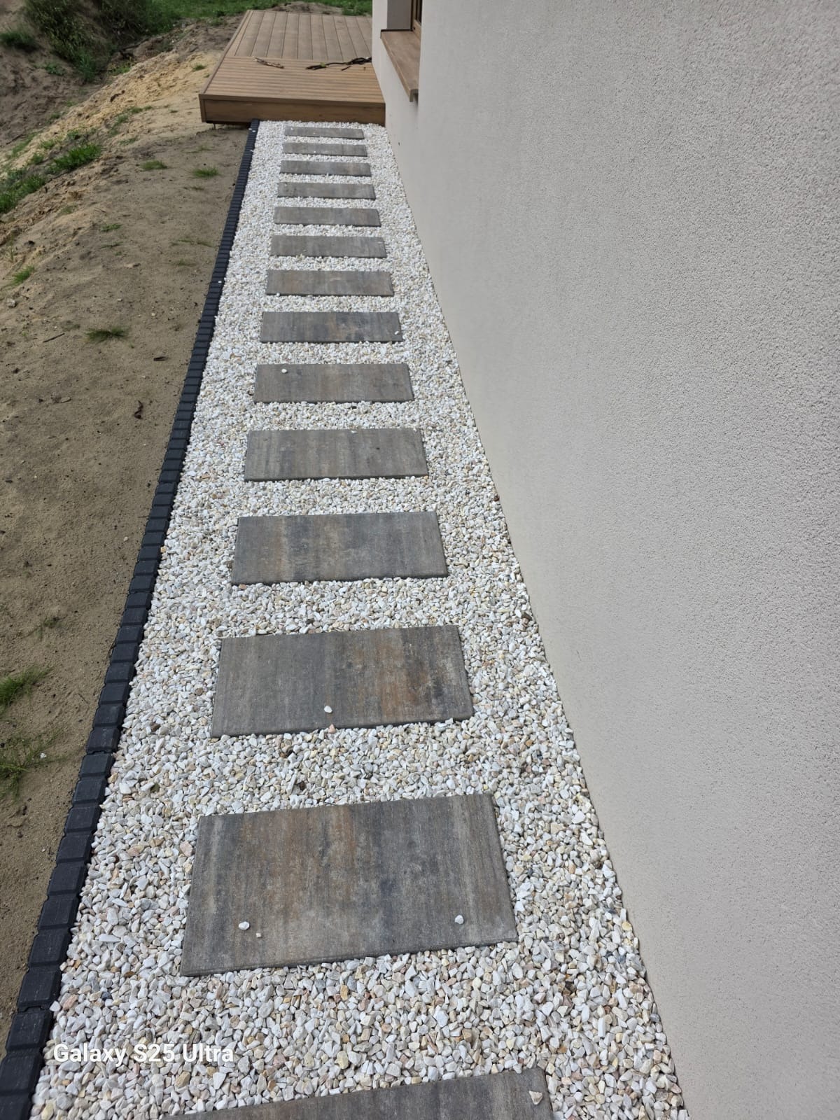 Ścieżka z płyt kamiennych ułożona w białym żwirze, obrzeże z czarnej kostki, widok z góry, przy budynku. Nowoczesne rozwiązanie aranżacji przestrzeni.