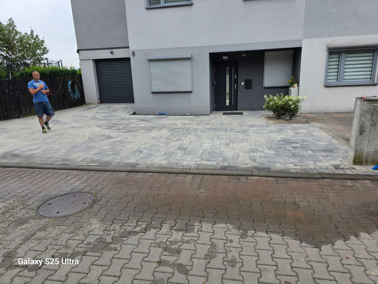 Nowoczesny podjazd z kostki brukowej przed domem z szarą elewacją i roletami. Widoczny fragment chodnika i studzienka kanalizacyjna. Osoba w tle.