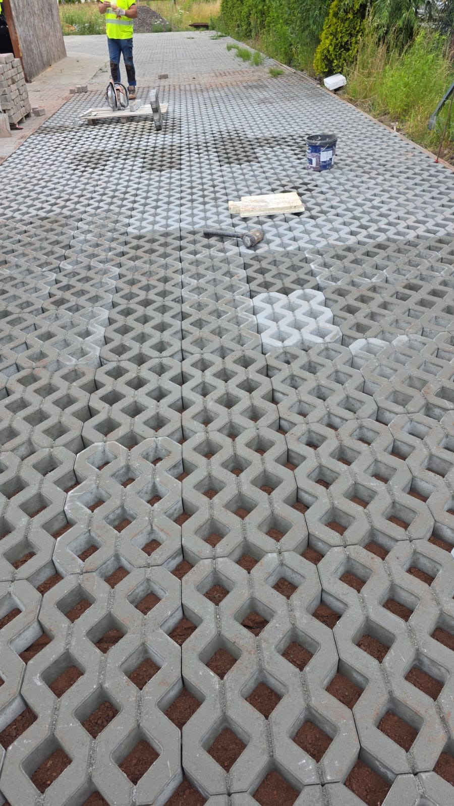Układanie ażurowej kostki brukowej na podsypce piaskowej. Fragment ułożonej nawierzchni, narzędzia i pracownik w odblaskowej kamizelce w tle.