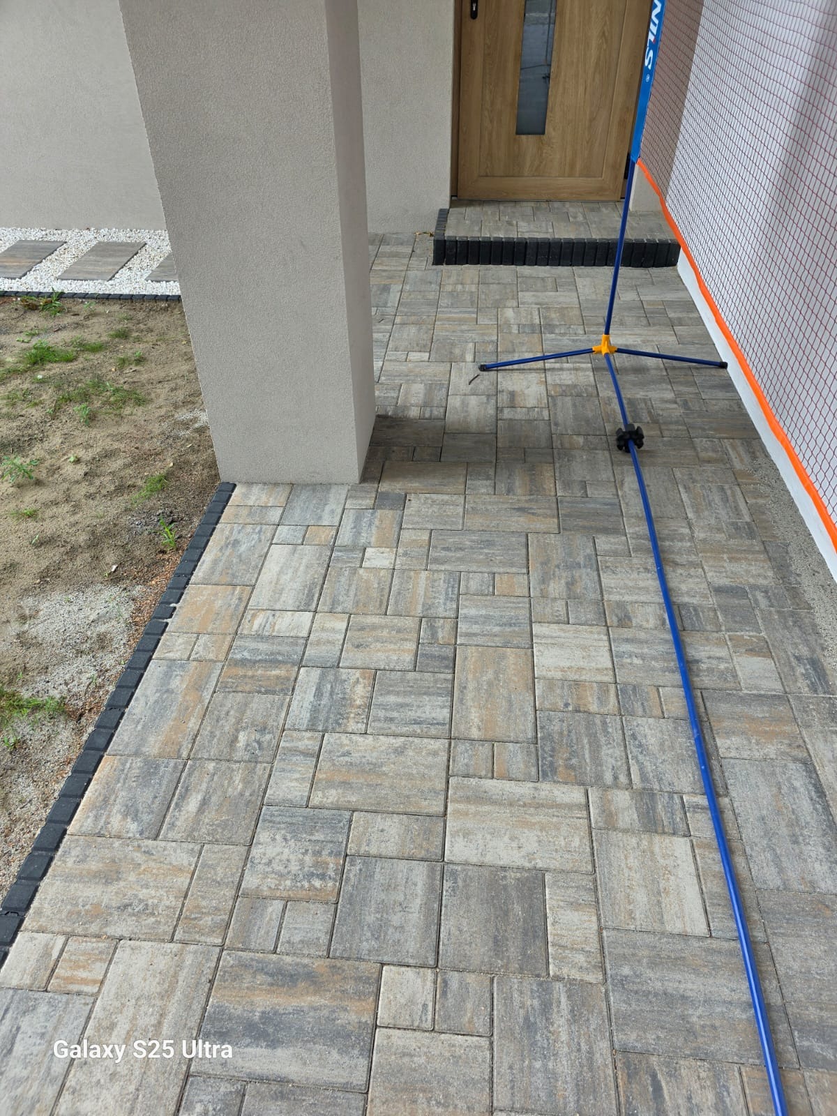 Ułożona kostka brukowa w odcieniach szarości i beżu tworzy wzór na podjeździe. Widoczny fragment słupka i drzwi. W tle siatka i rozłożony statyw.