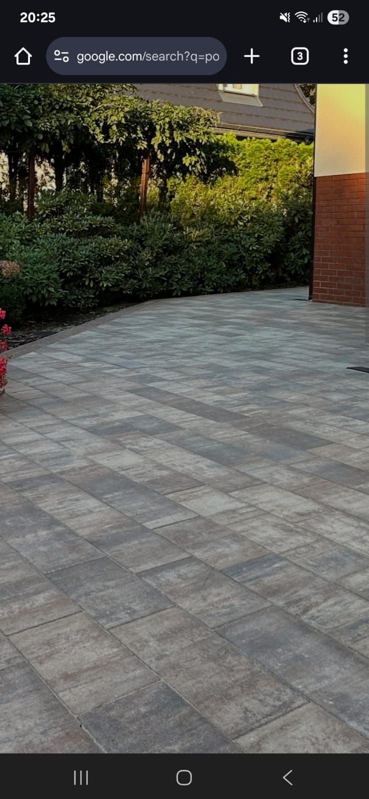 Ułożona kostka brukowa w odcieniach szarości na podjeździe, z zielenią w tle i fragmentem ceglanego muru, tworzy estetyczną przestrzeń przed domem.