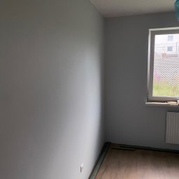 ROBOTA & PLUS - Szary pokój po malowaniu ścian z fragmentem drewnianej podłogi i listwami przypodłogowymi. Widoczne okno z widokiem na zewnątrz i grzejnik.