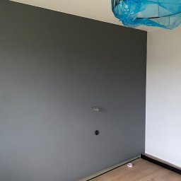 ROBOTA & PLUS - Ściana pomalowana na szaro z widocznymi gniazdkami, podłoga z paneli. Lampa zabezpieczona folią malarską. Widoczny fragment listwy przypodłogowej i taśmy malarskiej.