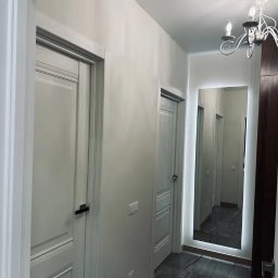 ROBOTA & PLUS - Korytarz po remoncie z białymi drzwiami, szarą marmurową podłogą i lustrem z podświetleniem LED. Widoczny żyrandol. Nowoczesny design.