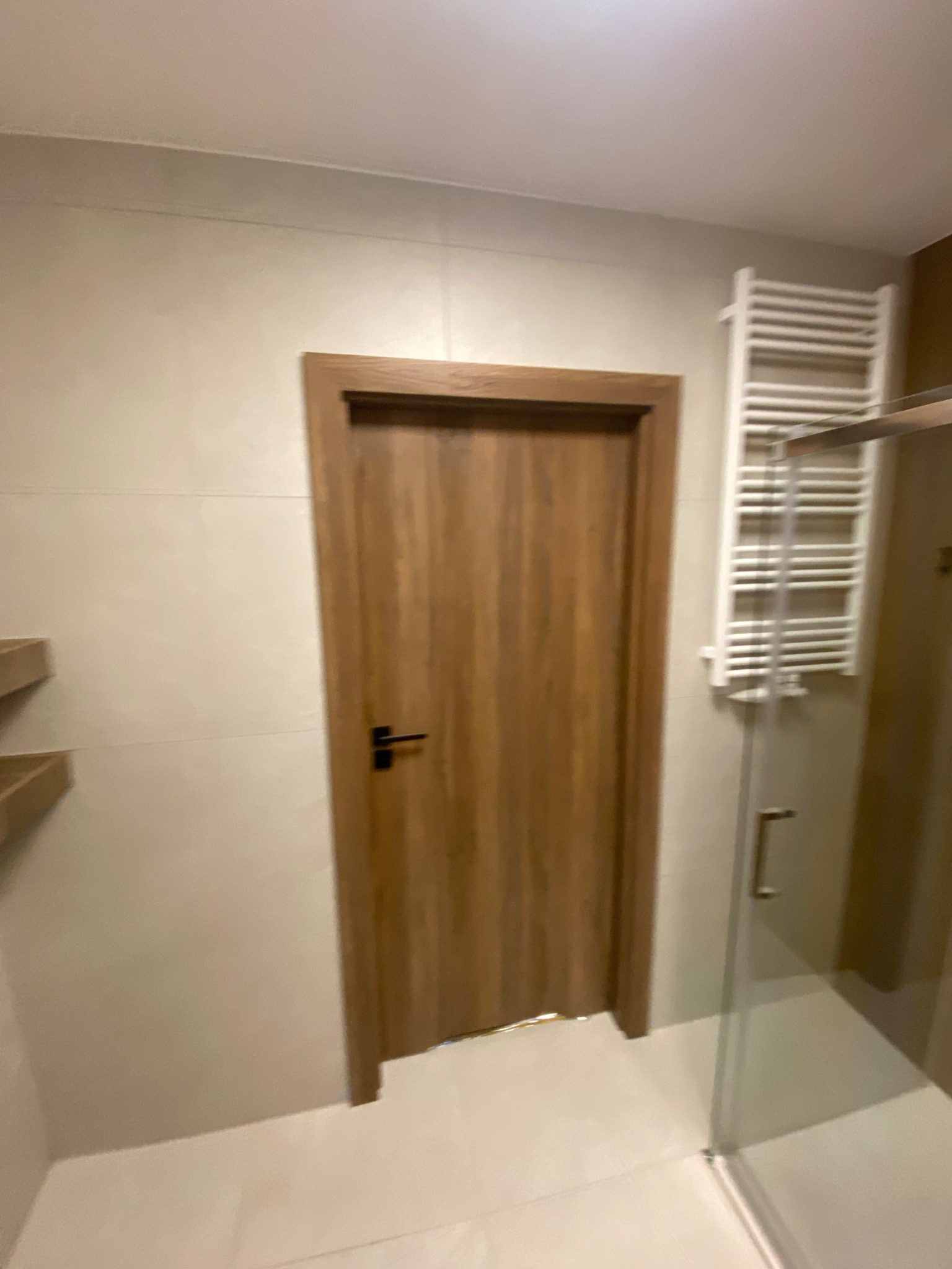 Wykończona łazienka z drewnianymi drzwiami i białym grzejnikiem drabinkowym. Jasne płytki na ścianach i podłodze. Fragment kabiny prysznicowej.