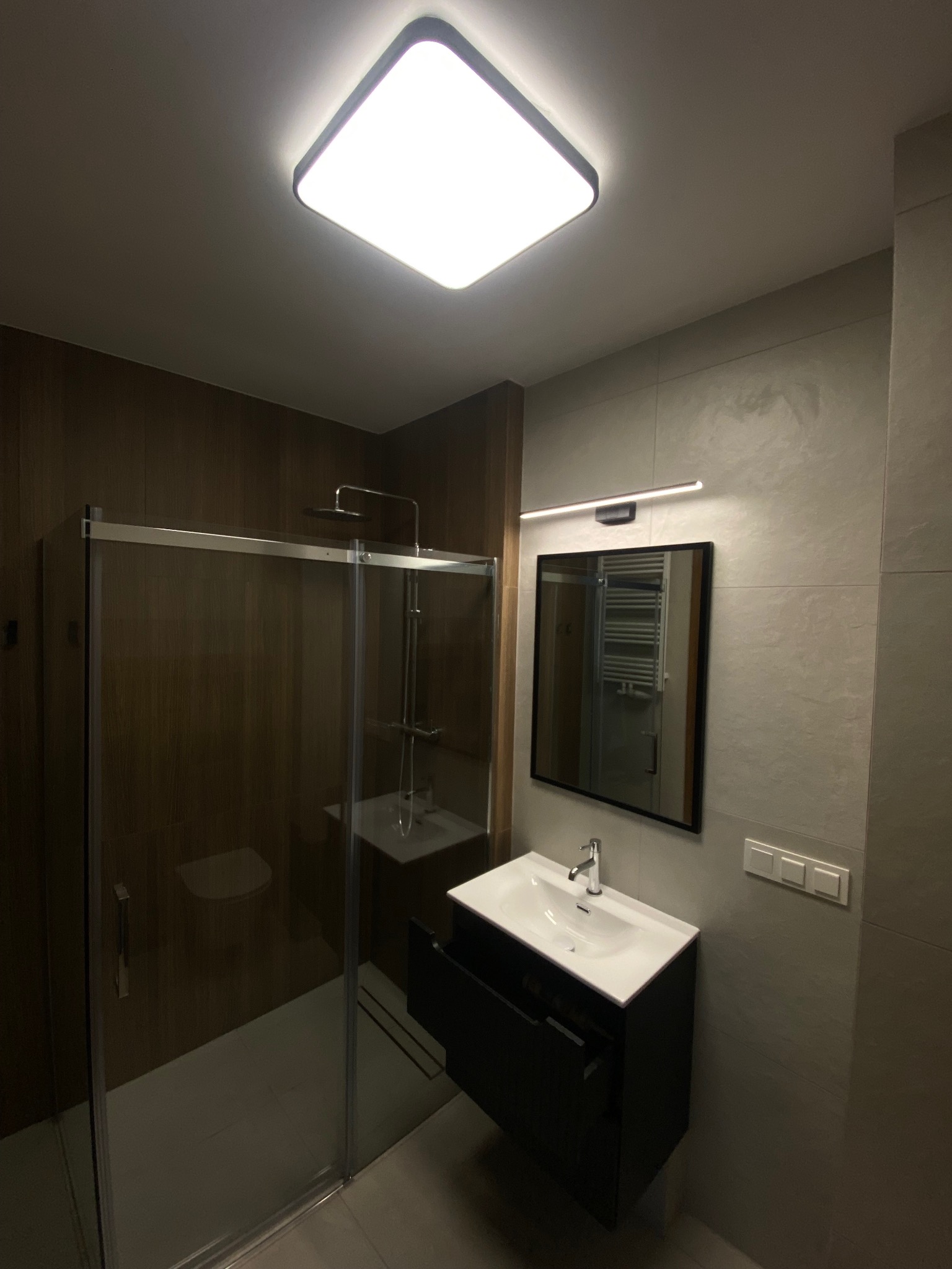 Nowoczesna łazienka z kabiną prysznicową typu walk-in, umywalką z szafką w kolorze czarnym i dużym lustrem z oświetleniem LED. Wykończenie w szarościach i brązach.