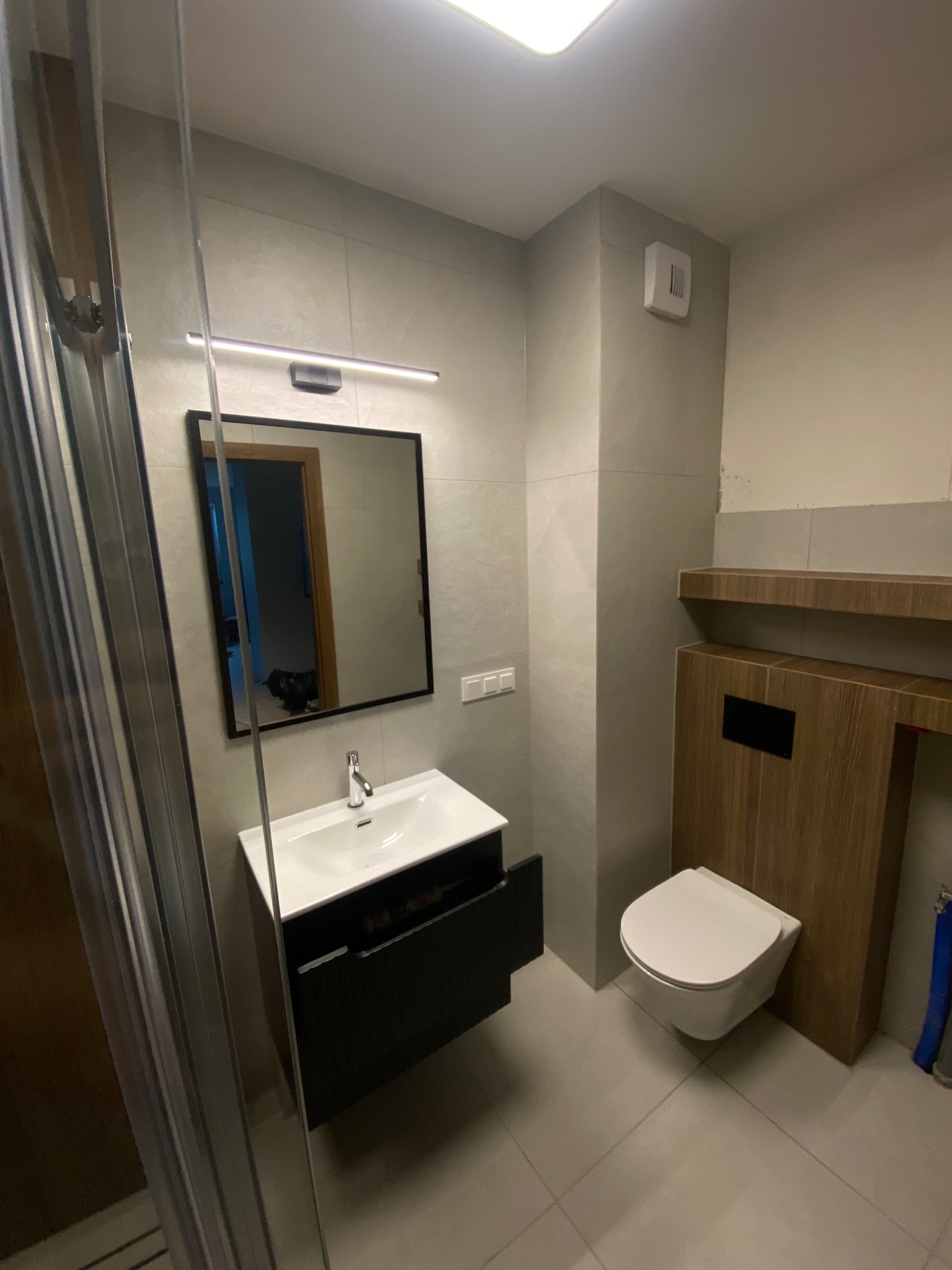 Nowoczesna łazienka z umywalką z szafką, lustrem podświetlanym LED i podwieszaną toaletą, wykończona w szarościach z akcentami drewna.