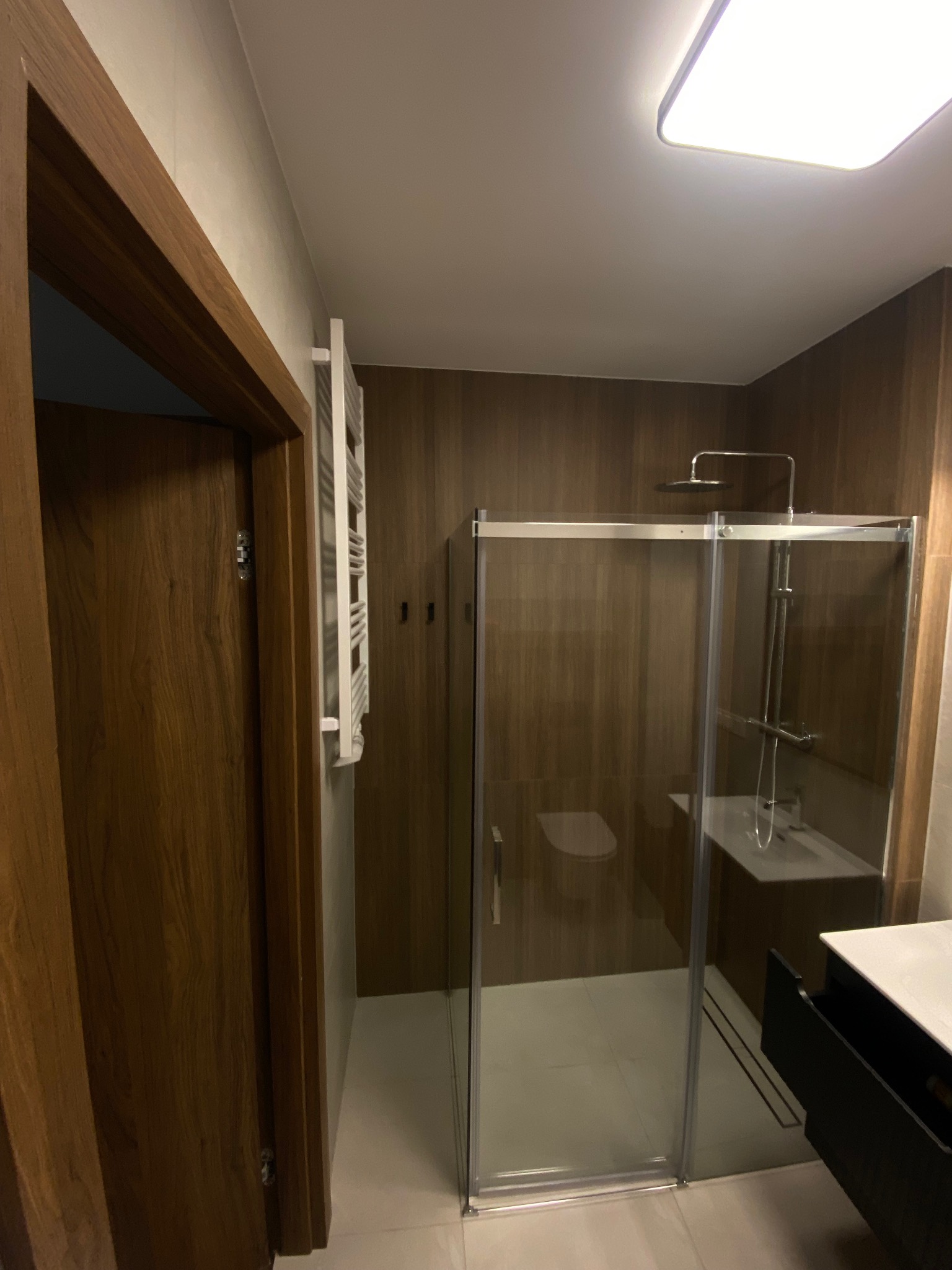 Nowoczesna łazienka z kabiną prysznicową typu walk-in, drewnopodobnymi panelami ściennymi i grzejnikiem drabinkowym. Minimalistyczny design, stonowana kolorystyka.