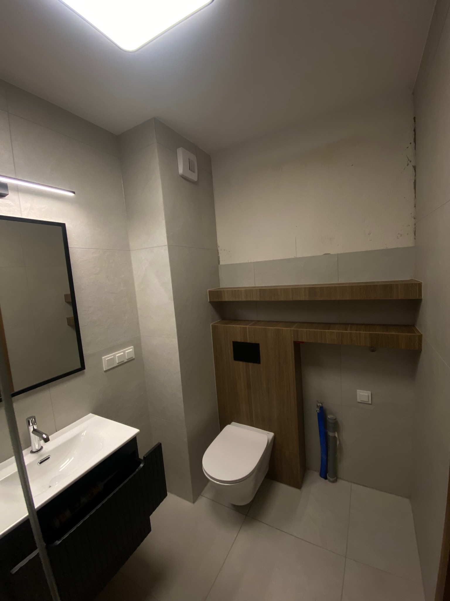 Nowoczesna łazienka z podwieszaną toaletą, umywalką z szafką w kolorze czarnym i półkami z drewnopodobnego materiału. Szare płytki na ścianach i podłodze.