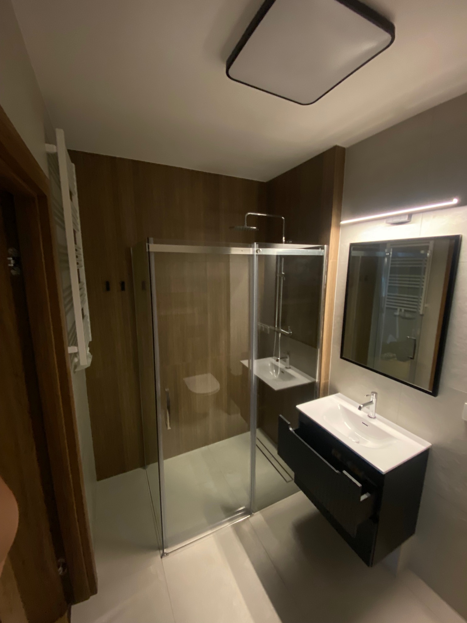 Nowoczesna łazienka po remoncie: kabina prysznicowa ze szklaną ścianą, umywalka z szafką w kolorze antracytowym, lustro z oświetleniem LED. Drewnopodobne wykończenie ściany.