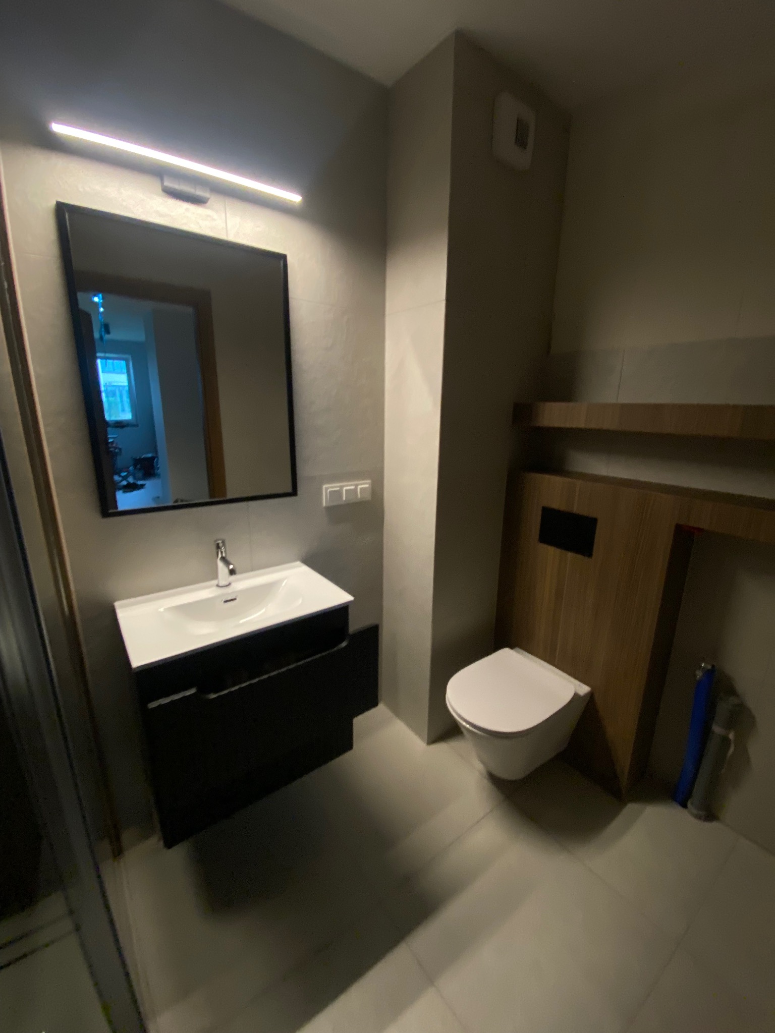 Nowoczesna łazienka z podwieszaną toaletą i czarną szafką pod umywalką, oświetlona ledowym światłem. Minimalistyczny design z drewnianymi akcentami.