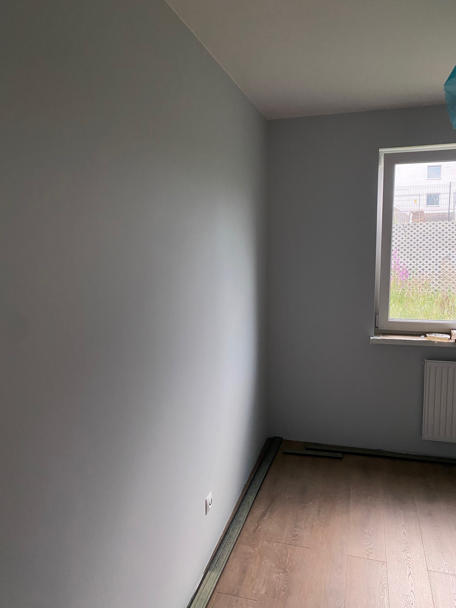 Szary pokój po malowaniu ścian z fragmentem drewnianej podłogi i listwami przypodłogowymi. Widoczne okno z widokiem na zewnątrz i grzejnik.