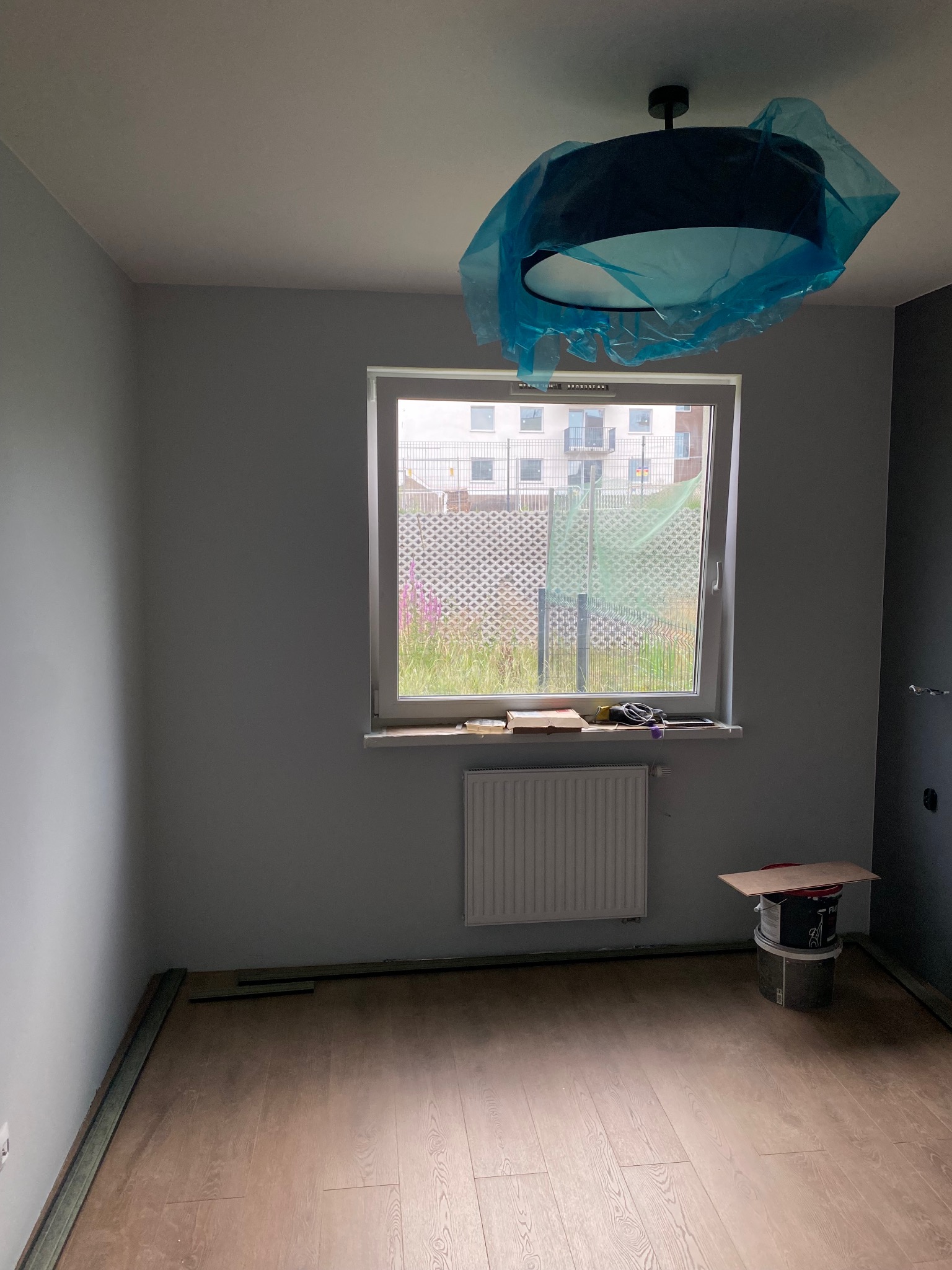 Pokój w trakcie remontu: panele podłogowe, grzejnik, okno z widokiem na zewnątrz, lampa zabezpieczona folią. Ściany pomalowane na szaro.