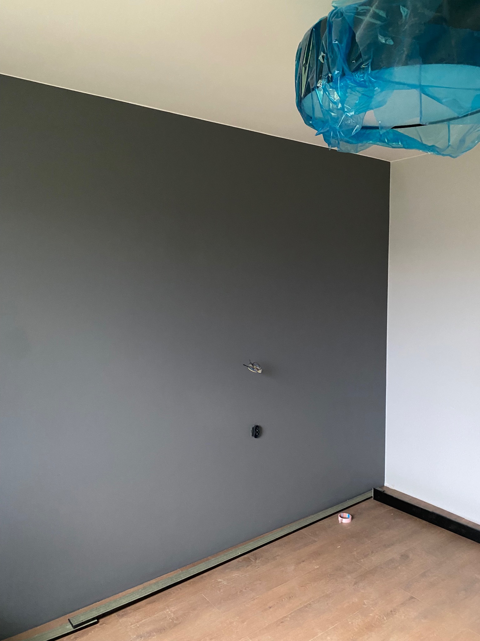 Ściana pomalowana na szaro z widocznymi gniazdkami, podłoga z paneli. Lampa zabezpieczona folią malarską. Widoczny fragment listwy przypodłogowej i taśmy malarskiej.