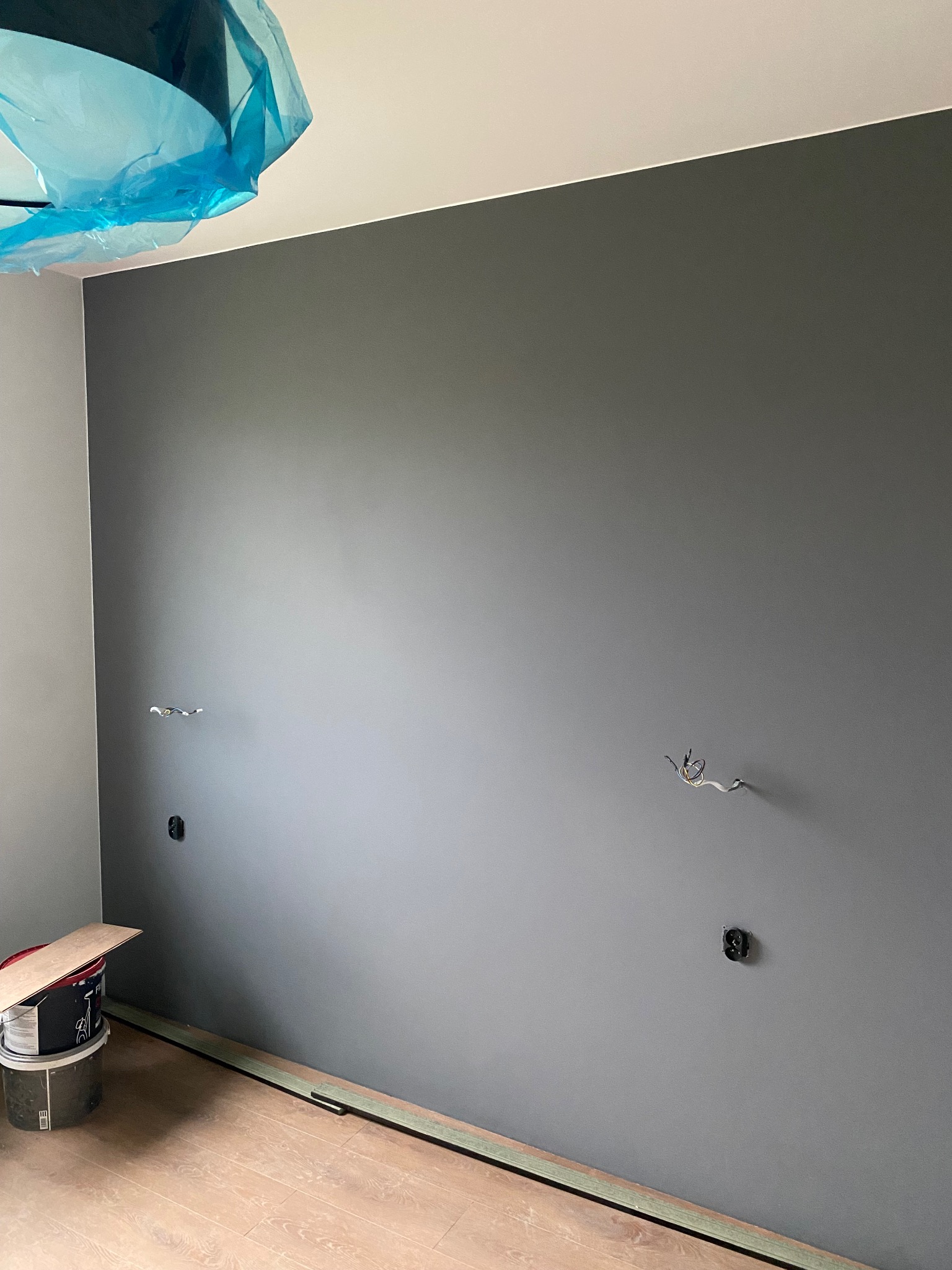 Ściana pomalowana na szaro z wyprowadzonymi przewodami elektrycznymi i gniazdkami. Na podłodze panele i wiadro z farbą. Sufit zabezpieczony folią malarską.