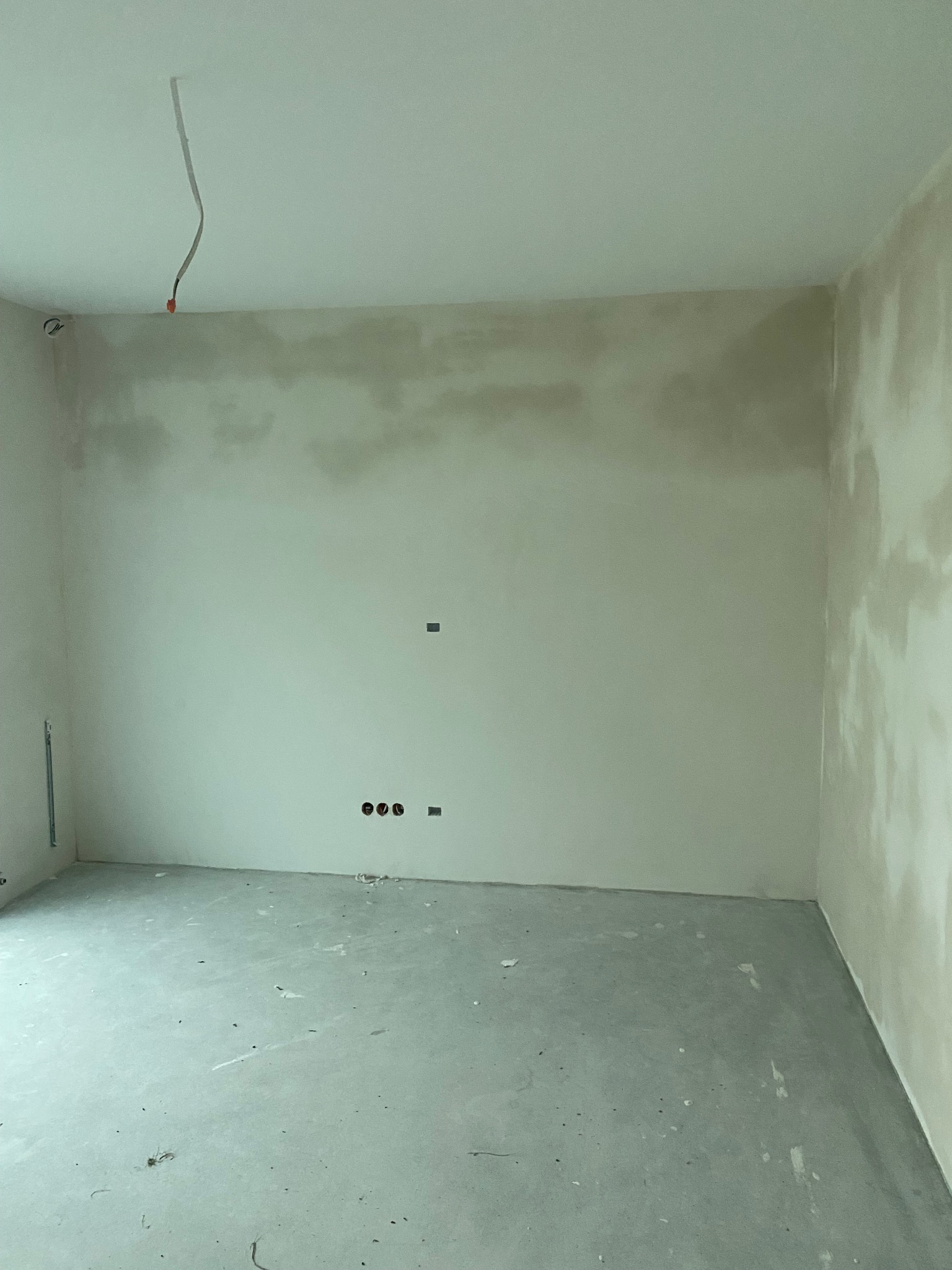 Surowe wnętrze w trakcie prac wykończeniowych: ściany po szpachlowaniu, widoczne kable i puszki elektryczne. Podłoga betonowa, przygotowana do dalszych prac.