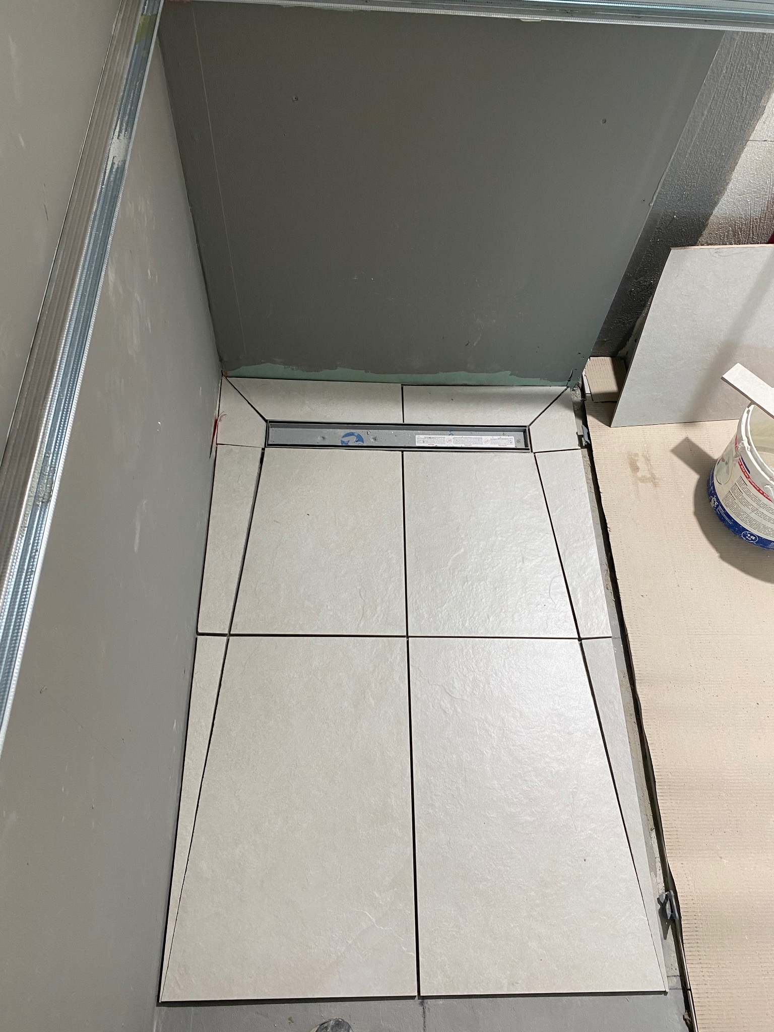 Wykończenie posadzki w kabinie prysznicowej z odpływem liniowym. Jasne, kwadratowe płytki ułożone na podłodze, widoczne elementy konstrukcji ścian.