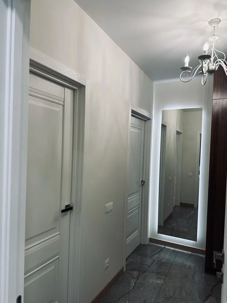 Korytarz po remoncie z białymi drzwiami, szarą marmurową podłogą i lustrem z podświetleniem LED. Widoczny żyrandol. Nowoczesny design.