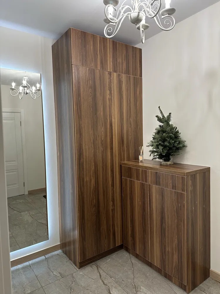 Szafa wnękowa z lustrem w przedpokoju, wykończona okleiną w kolorze ciemnego drewna. Oświetlenie LED wokół lustra i marmurowe płytki podłogowe.