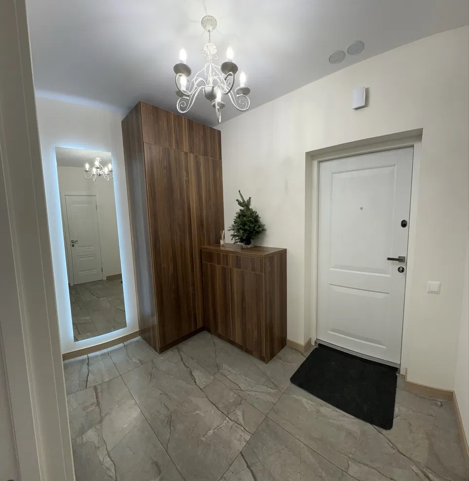 Nowoczesny przedpokój: szafa z drewnianymi frontami, lustro z podświetleniem LED, szare płytki podłogowe i białe drzwi wejściowe z czarną wycieraczką. Minimalistyczny design.