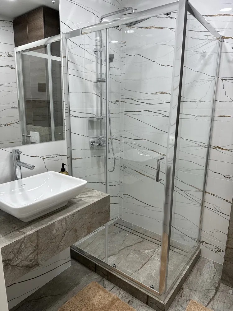 Nowoczesna kabina prysznicowa w łazience z marmurowymi płytkami, umywalką nablatową i lustrem. Chromowane wykończenia.