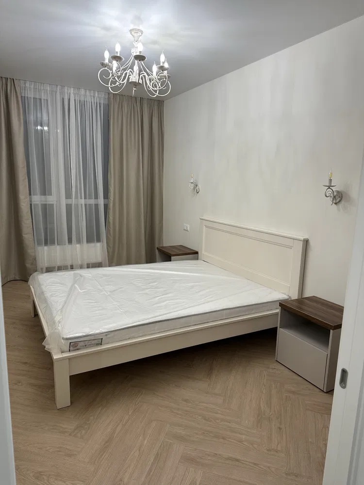 Jasna sypialnia z parkietem w jodełkę, białym łóżkiem z materacem w folii, szafkami nocnymi i żyrandolem. Minimalistyczny design, świeżo po remoncie.