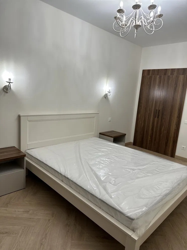 Jasne wnętrze sypialni z łóżkiem w folii, szafkami nocnymi i parkietem ułożonym w jodełkę. Na ścianie kinkiety, a z sufitu zwisa ozdobny, biały żyrandol.