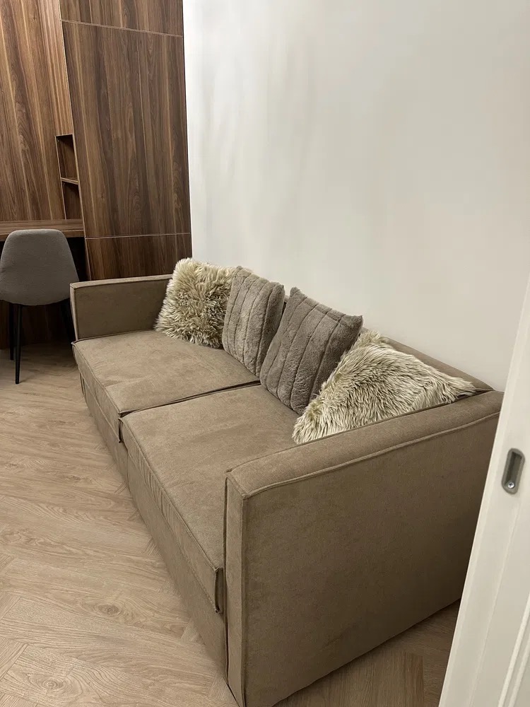 Beżowa sofa z poduszkami w pokoju z drewnianą zabudową biurka i jasną ścianą. Minimalistyczny wystrój wnętrza.