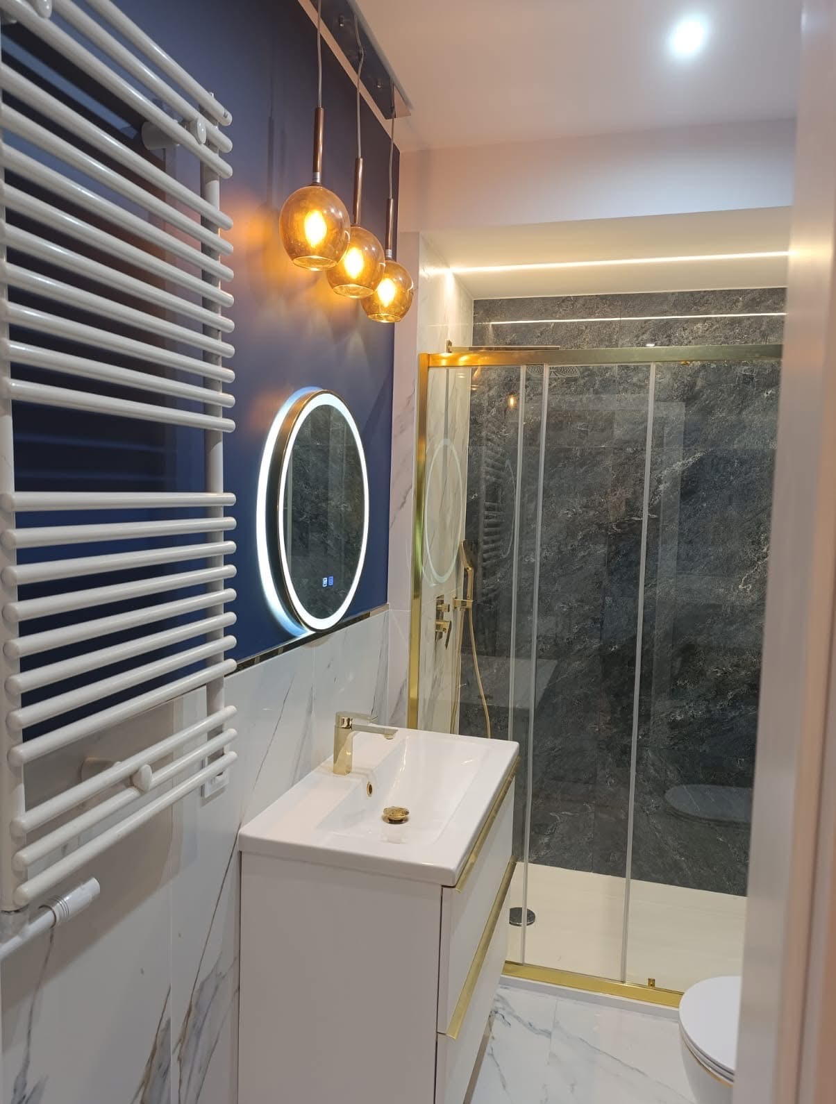 Elegancka łazienka z kabiną prysznicową, umywalką z szafką w bieli i złotych akcentach. Na ścianie podświetlane lustro i grzejnik drabinkowy. Oświetlenie punktowe.