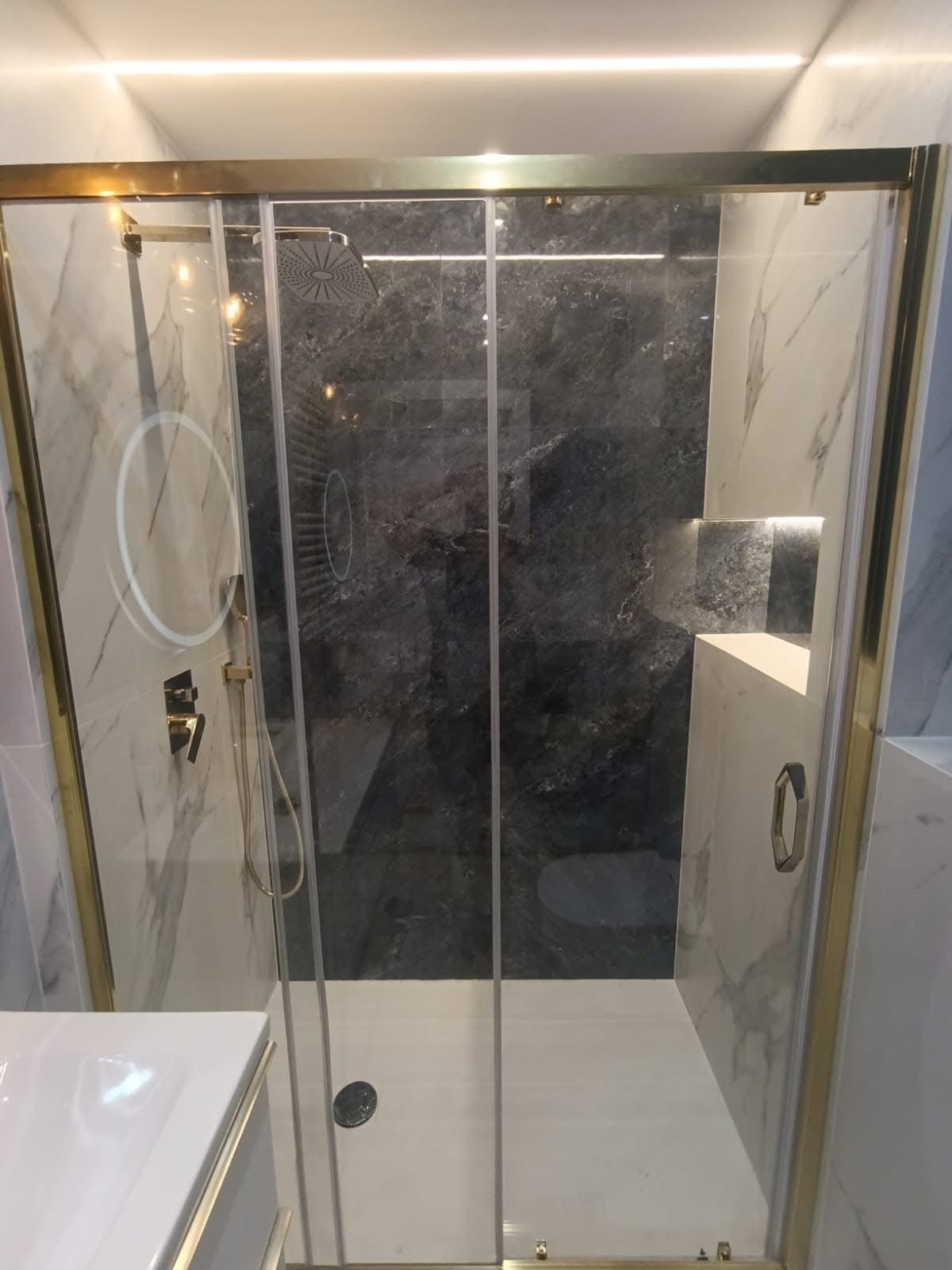 Nowoczesna kabina prysznicowa z marmurowymi ścianami, złotymi detalami i oświetleniem LED. Widoczna deszczownica i bateria.