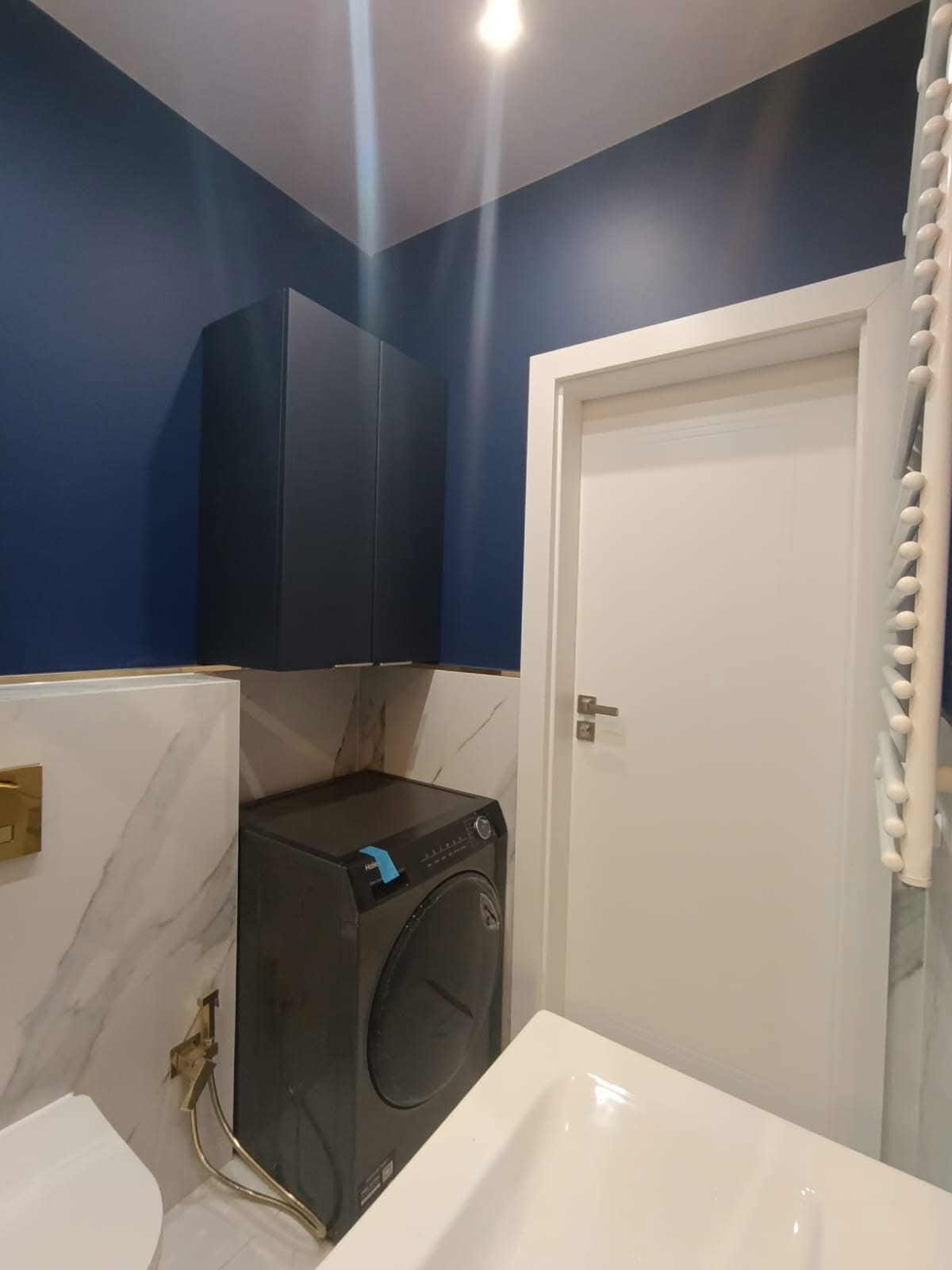 Nowoczesna łazienka po remoncie: granatowe ściany, marmurowe płytki, zabudowa nad pralką i biała armatura. Widoczne detale wykończenia.
