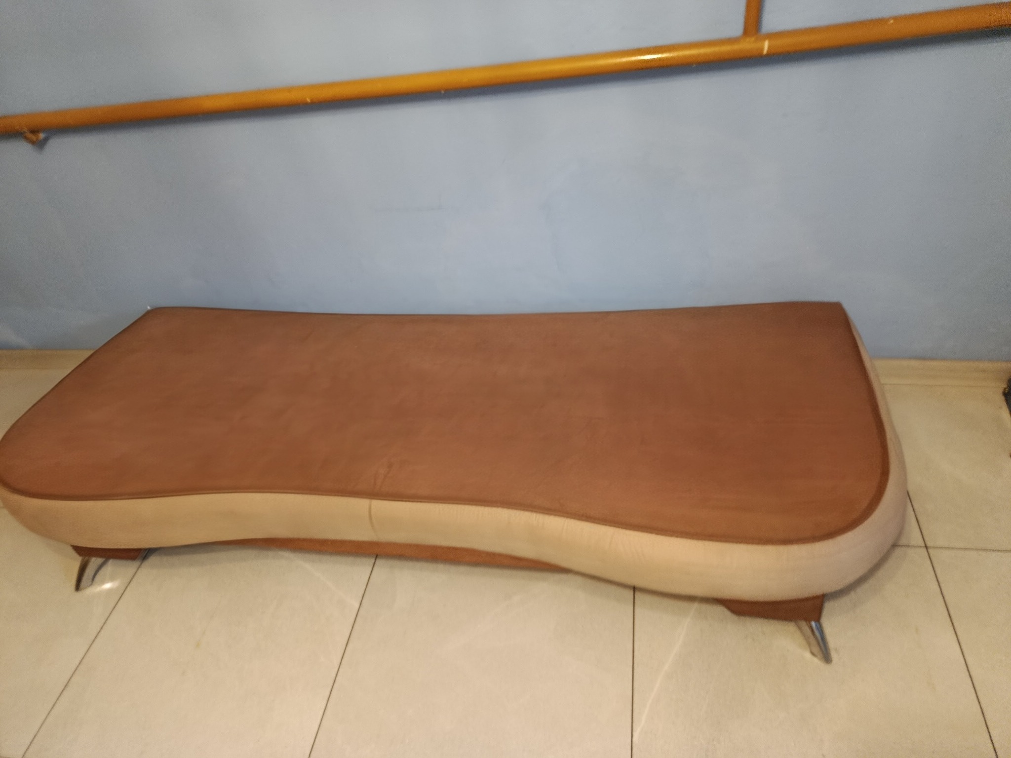 Brązowa tapicerowana pufa na metalowych nóżkach, stojąca na jasnych płytkach. U góry widoczny fragment poręczy. Stonowana kolorystyka, miękkie światło.