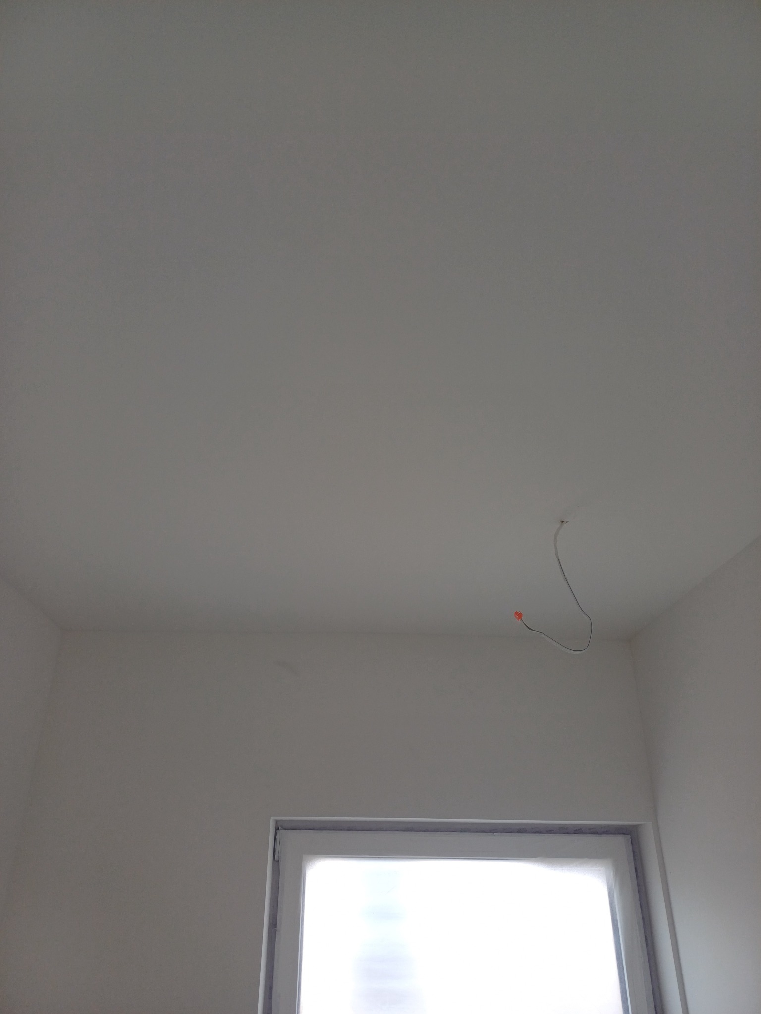 Białe, świeżo pomalowane wnętrze z widocznym kablem wystającym z sufitu i oknem w dolnej części kadru. Minimalistyczny, jasny pokój po remoncie.