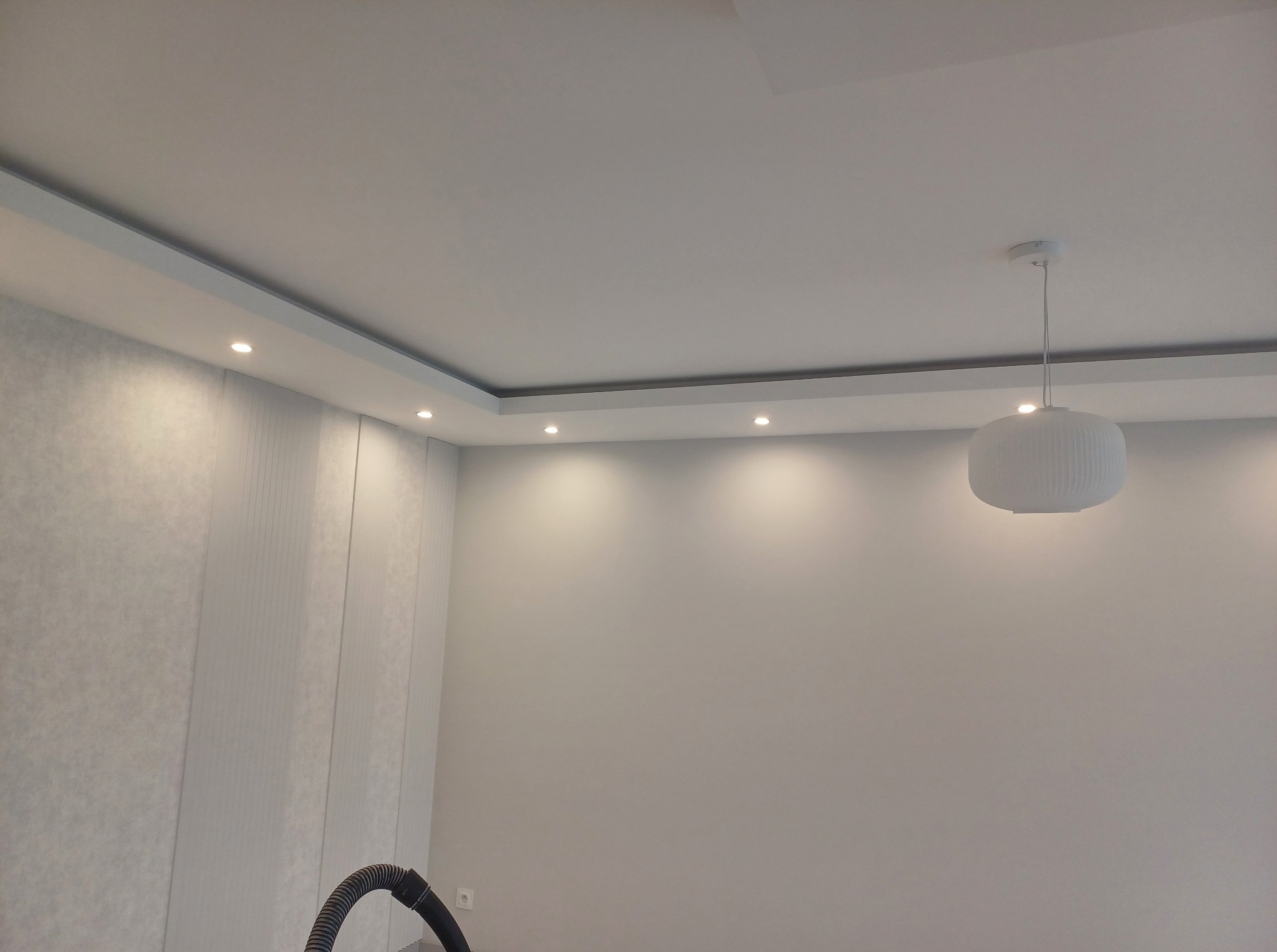 Nowoczesne wykończenie sufitu z oświetleniem punktowym i ozdobną listwą. Ściany wykończone w jasnych odcieniach, widoczna lampa wisząca i fragment odkurzacza.