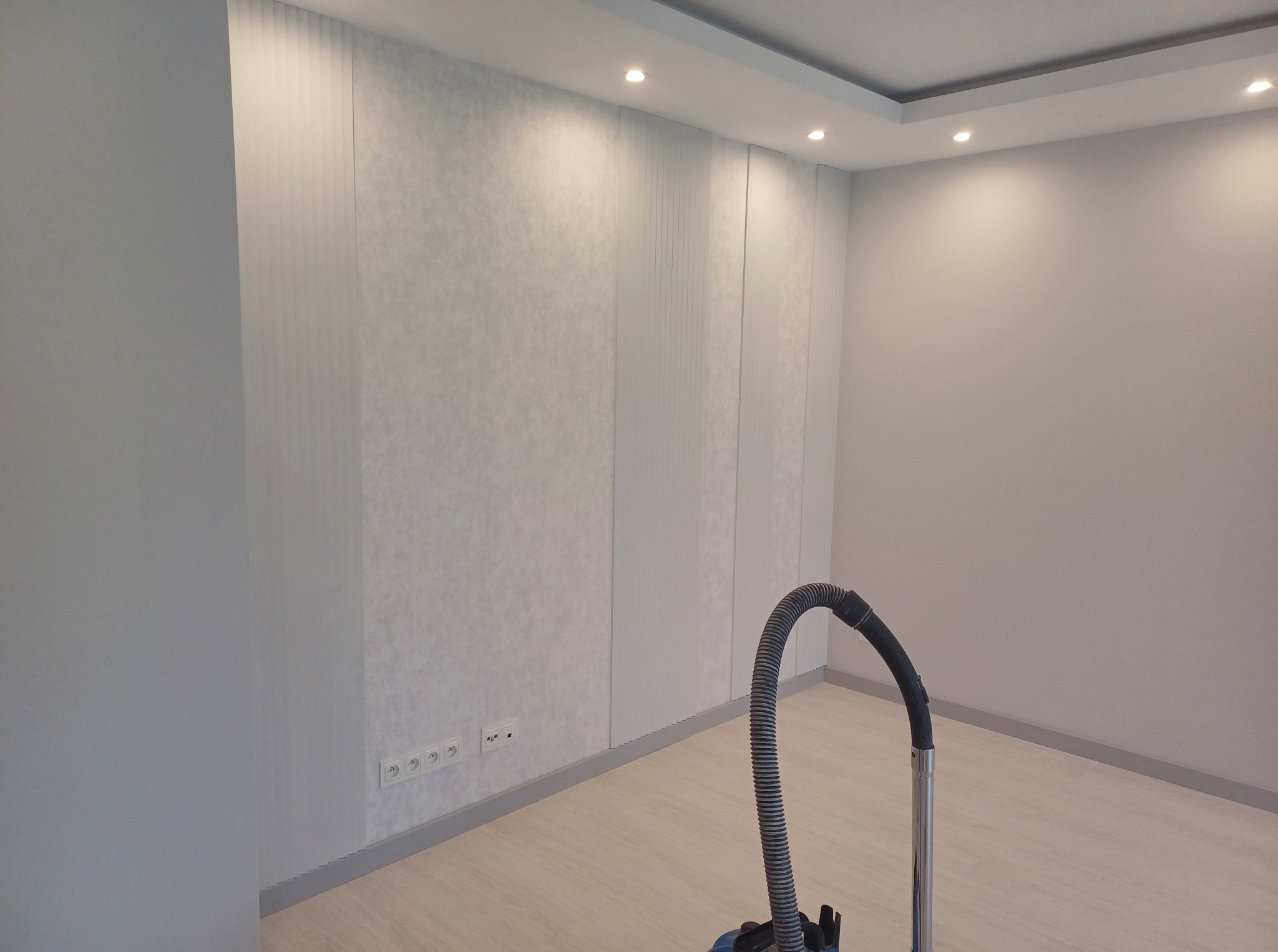 Wykończone wnętrze pokoju z panelami ściennymi i gładzią, widoczne gniazdka elektryczne przy podłodze, oświetlenie sufitowe i odkurzacz w rogu. Jasna kolorystyka.