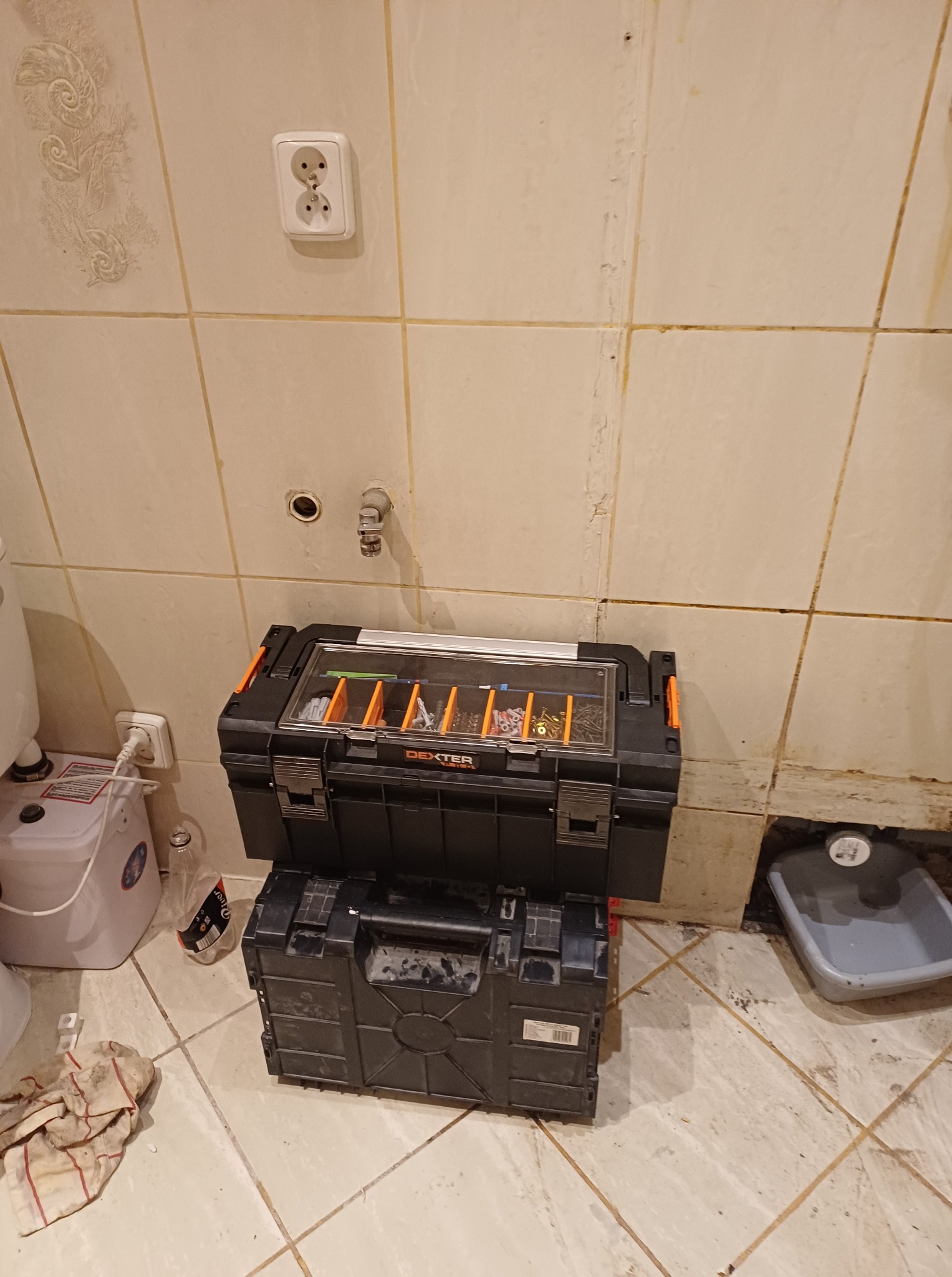 Dwie czarne skrzynki narzędziowe Dexter ustawione jedna na drugiej w łazience z beżowymi płytkami. Widoczne narzędzia w górnej skrzynce, gniazdko elektryczne i fragment instalacji wodnej.