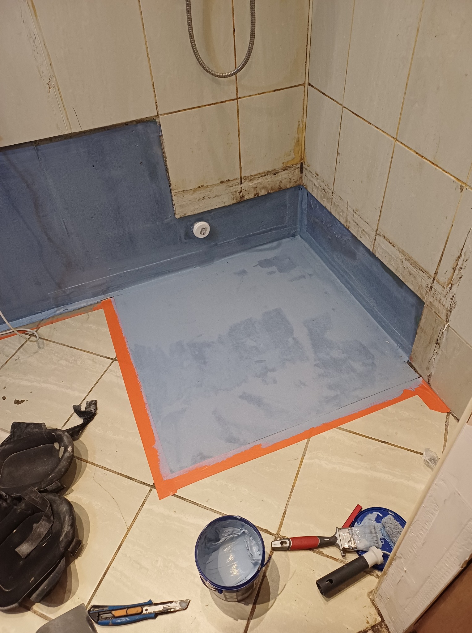 Hydroizolacja w narożniku łazienki, zabezpieczona taśmą malarską. Widoczne narzędzia: pędzle, wiadro z farbą, nożyk. Prace wykończeniowe w trakcie realizacji.