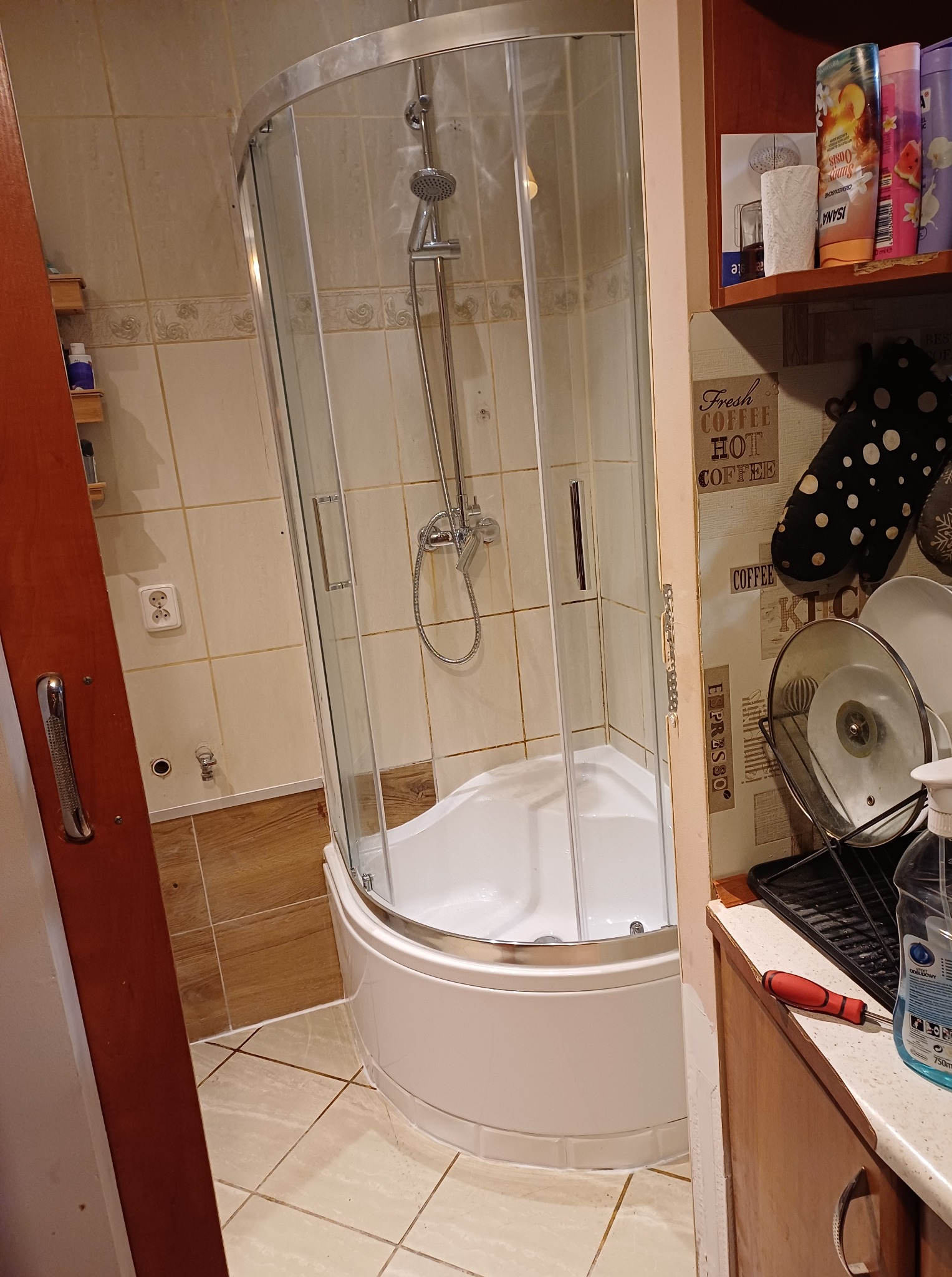 Narożna kabina prysznicowa z chromowaną armaturą i białym brodzikiem, zamontowana w łazience z beżowymi płytkami. Widoczny fragment szafki z akcesoriami.