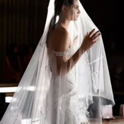 Panna młoda w profilu, w bogato zdobionej sukni ślubnej, z welonem zakrywającym twarz, dłonie uniesione w geście delikatności, na ciemnym tle.