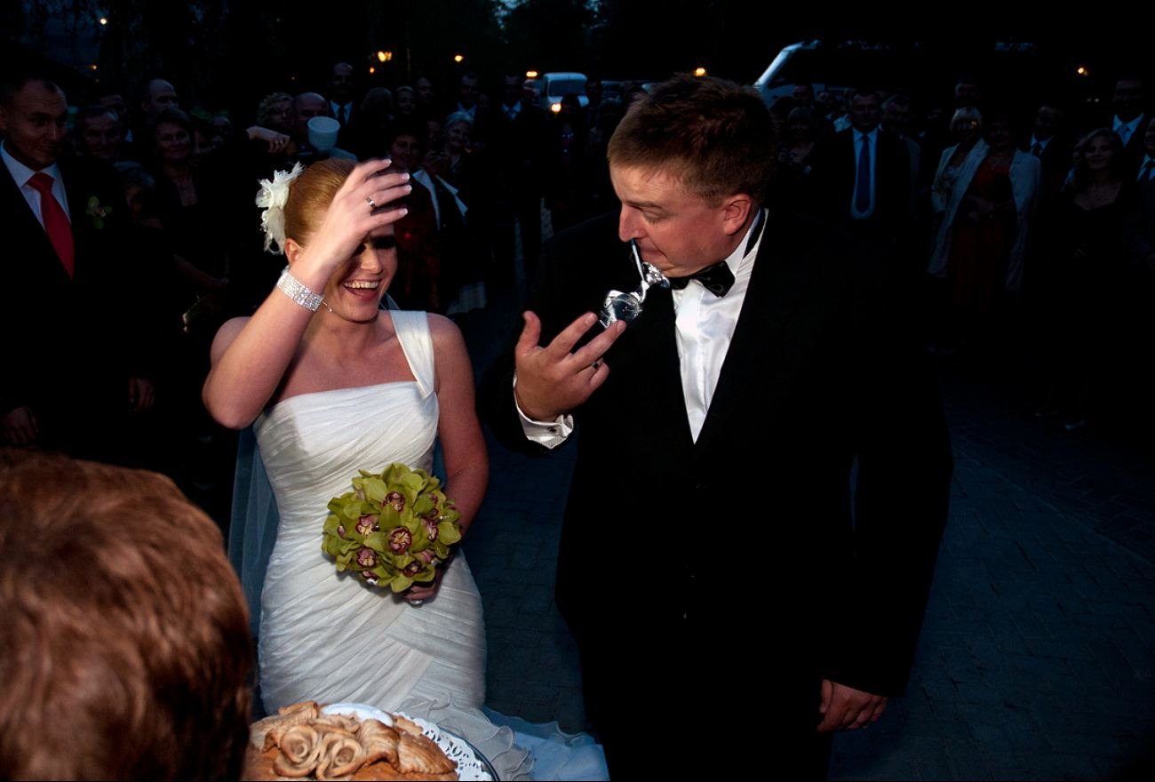 Uśmiechnięta panna młoda w białej sukni i pan młody w smokingu, wznoszący toast kieliszkiem na tle gości weselnych w plenerze po zmroku.