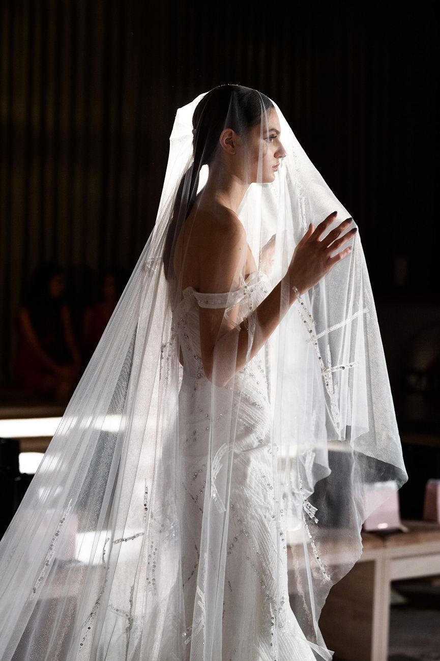 Panna młoda w profilu, w bogato zdobionej sukni ślubnej, z welonem zakrywającym twarz, dłonie uniesione w geście delikatności, na ciemnym tle.
