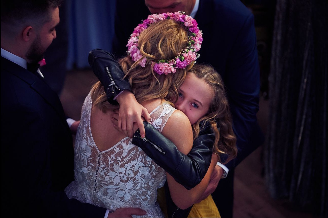 Panna młoda w koronie z kwiatów i koronkowej sukni obejmuje dziewczynkę w czarnej kurtce na tle mężczyzn w garniturach podczas uroczystości.