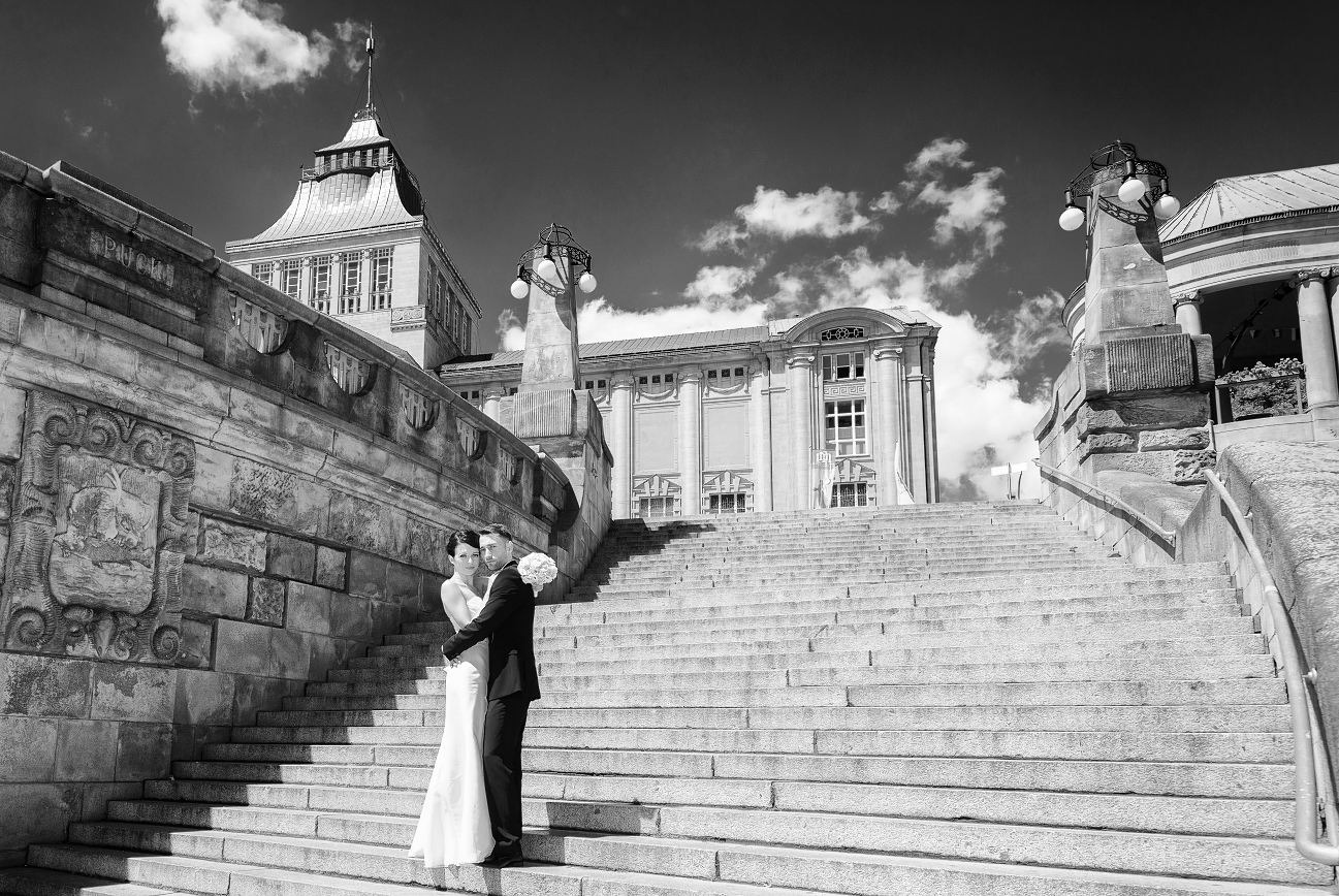 Czarno-białe zdjęcie pary młodej na tle monumentalnych schodów i architektury w stylu retro, z dekoracyjnymi lampami i rzeźbieniami na balustradach.