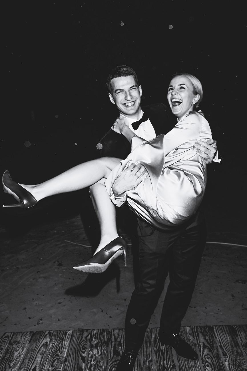 Czarno-białe zdjęcie uśmiechniętej pary: mężczyzna w smokingu trzyma na rękach śmiejącą się kobietę w sukience i szpilkach na tle ciemnego otoczenia.