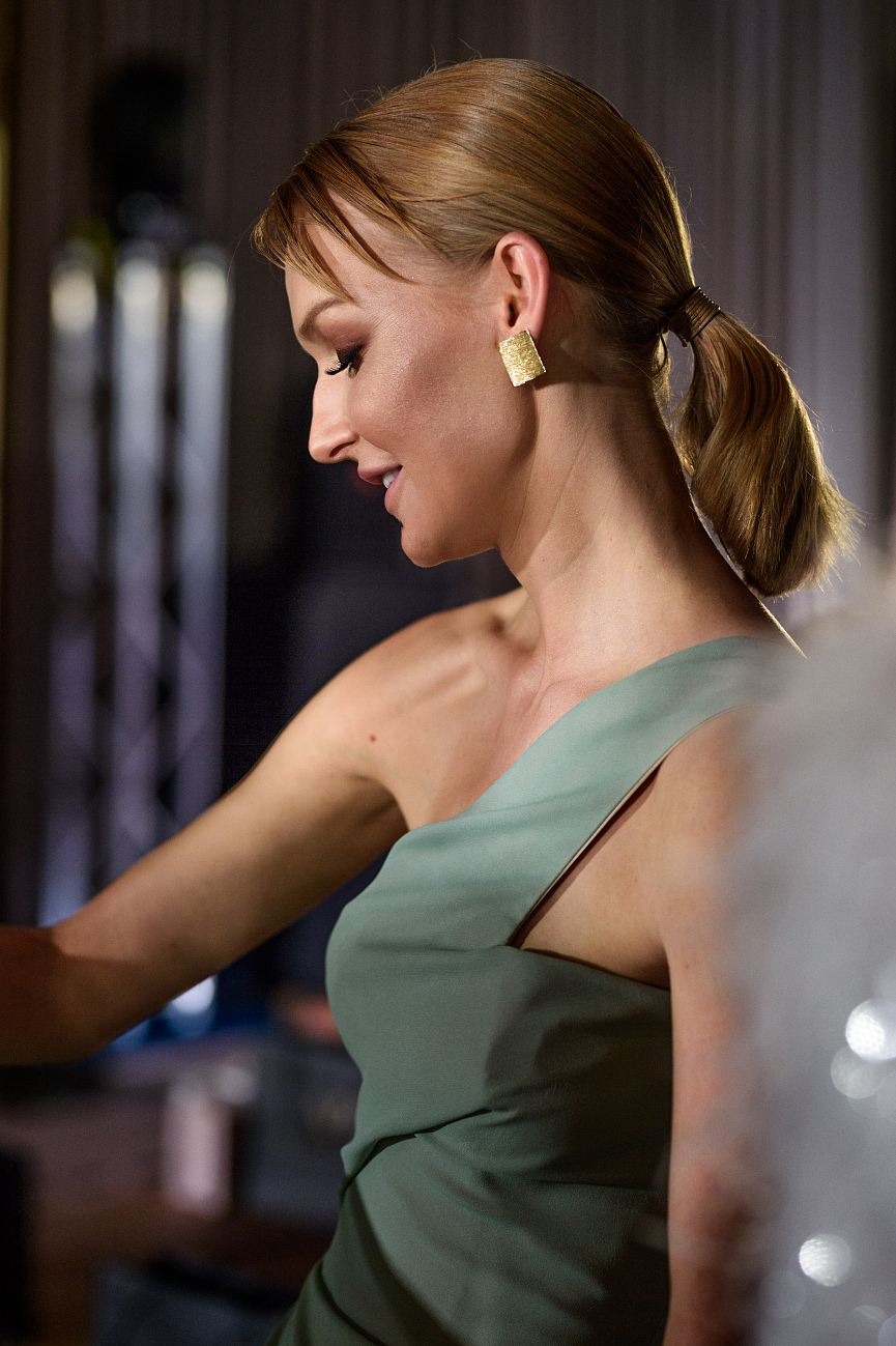 Portret kobiety z profilu w zielonej sukience i złotych kolczykach na tle ciemnego wnętrza. Ujęcie z bliska, skupiające się na detalu biżuterii i eleganckiej stylizacji.