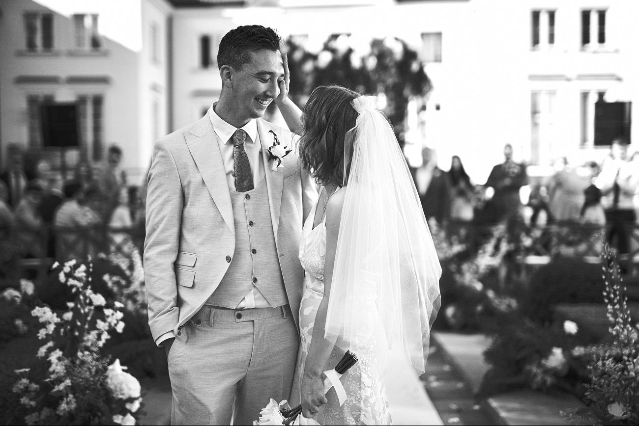 Czarno-białe zdjęcie uśmiechniętej pary młodej w jasnych strojach, z gośćmi w tle, na tle jasnego budynku z oknami. Pan młody trzyma rękę w kieszeni.