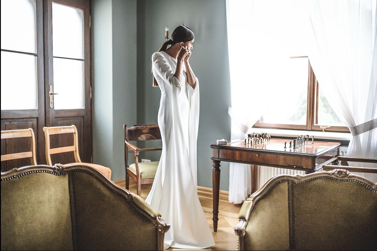 Panna młoda w białej sukni, poprawia kolczyk w uchu, stojąc w pokoju z szachami na stole. Ujęcie z sesji ślubnej, w tle okno z białymi zasłonami.